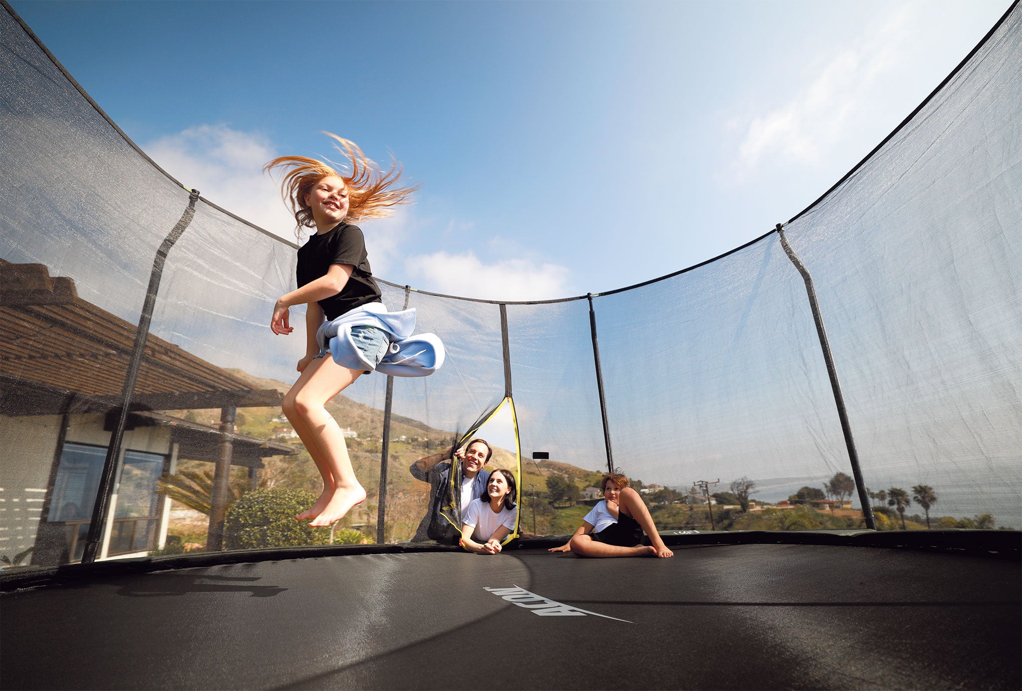 Tyttö hyppii ACON trampoliinilla, johon on asennettuna premium tason turvaverkko. Muut lapset katsovat pomppivaa tyttöä.