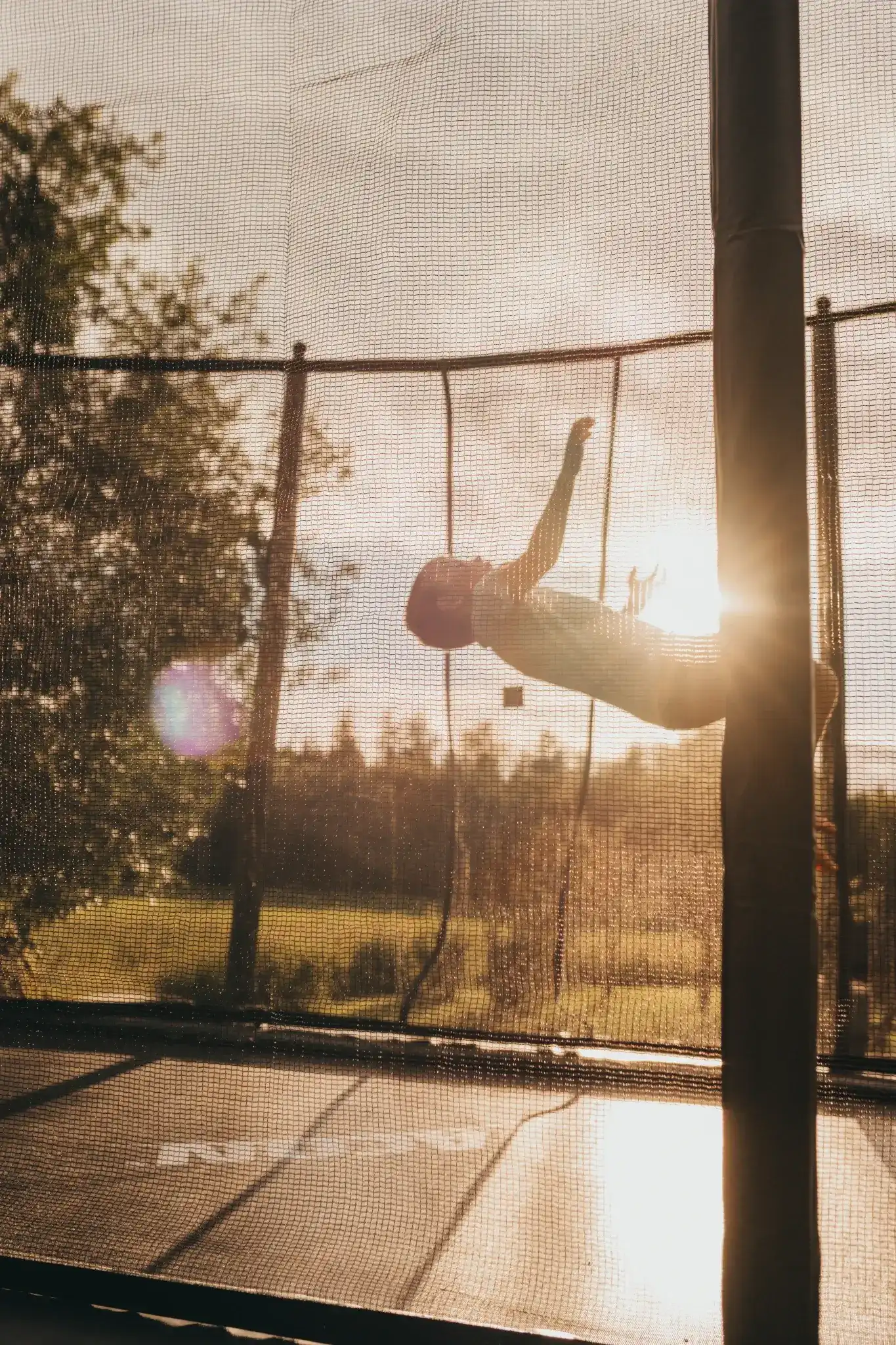 Lapsi pomppii trampoliinilla ilta-auringossa, valo heijastuu kauniisti trampoliinin suojaverkon läpi.