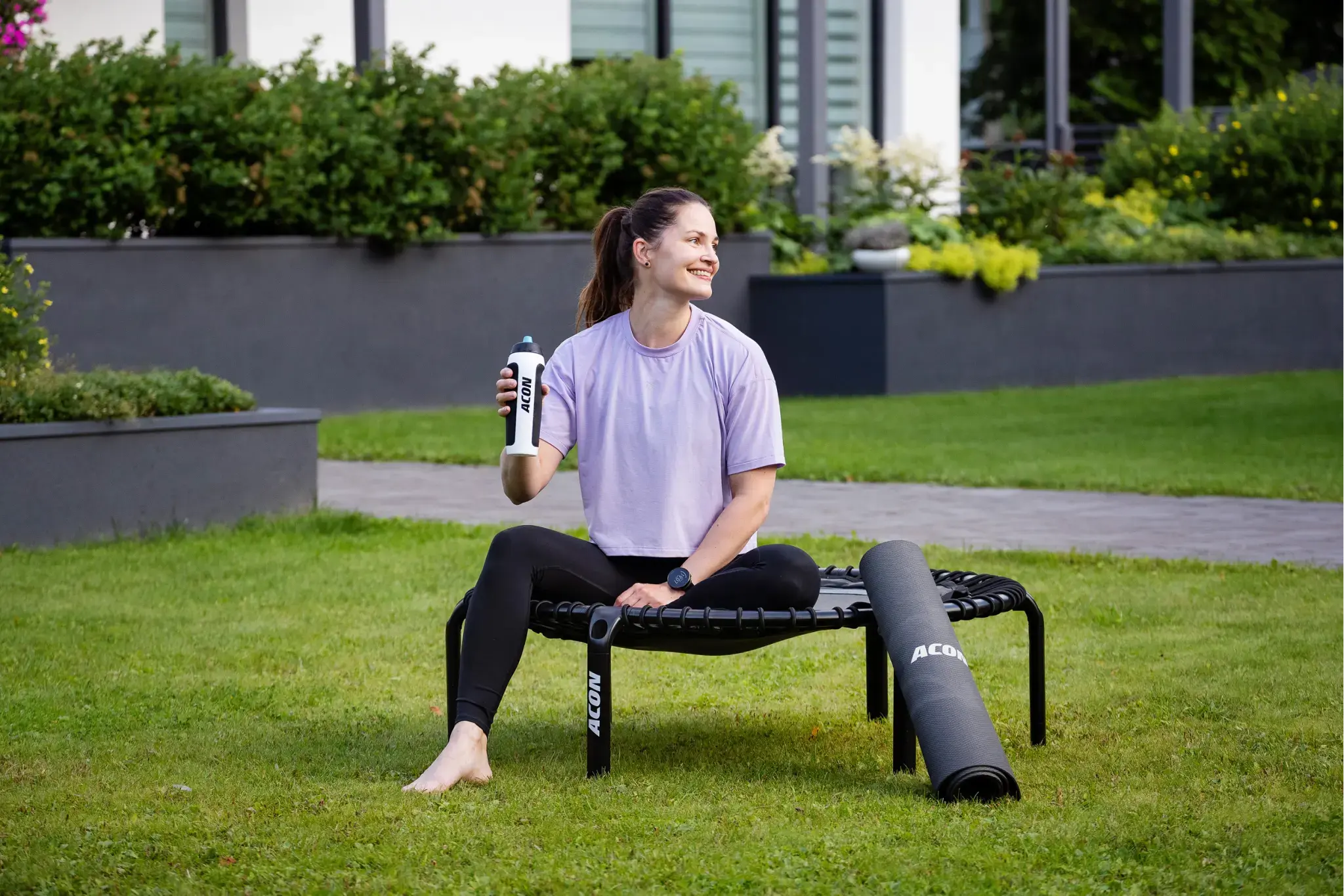 Nainen violetissa paidassa istuu ulkona Acon fitness-trampoliinilla Acon-juomapullon ja jumppamaton kera.