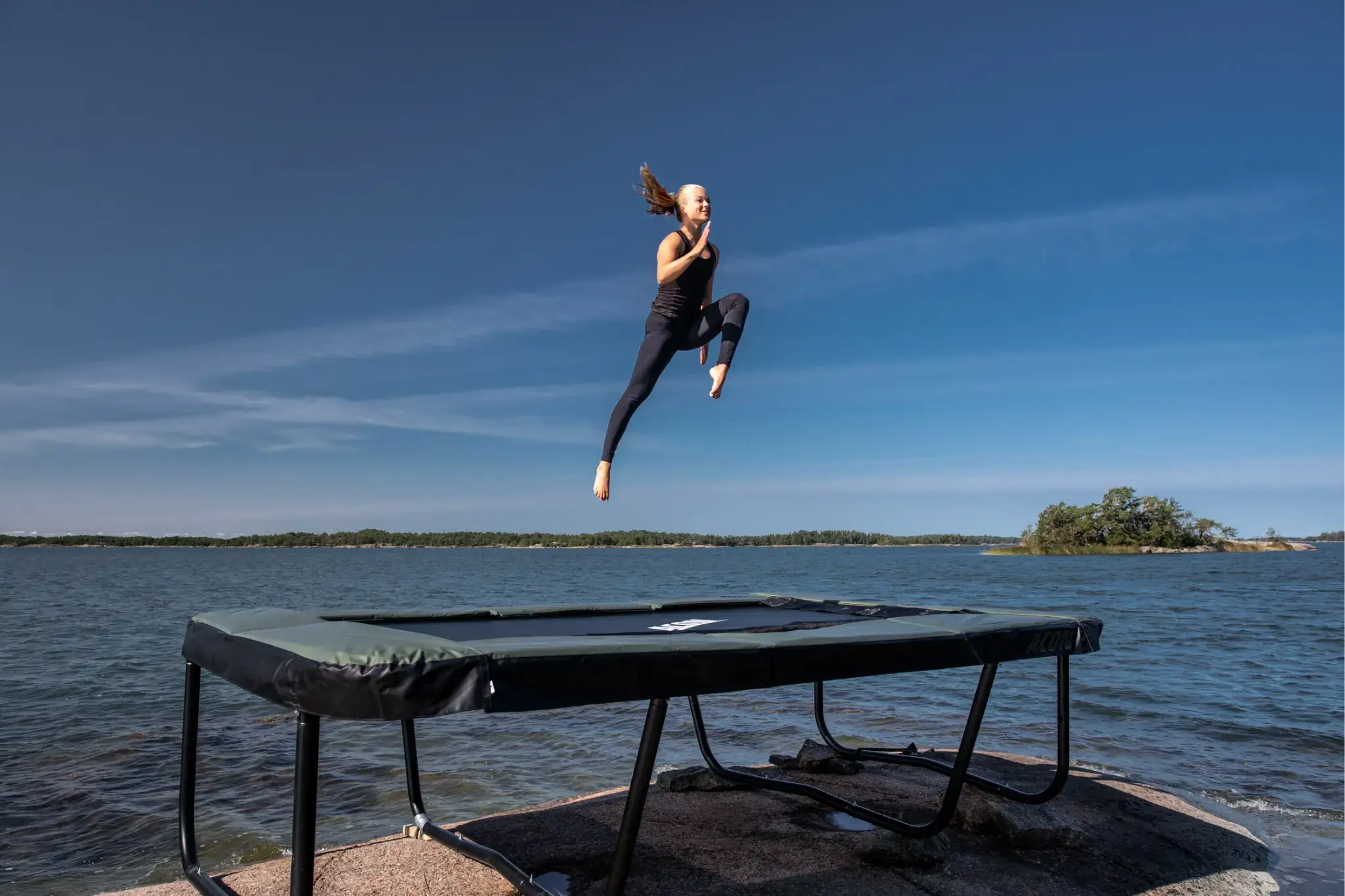 Nainen hyppäämässää korkealle Acon trampoliinilla veden äärellä.