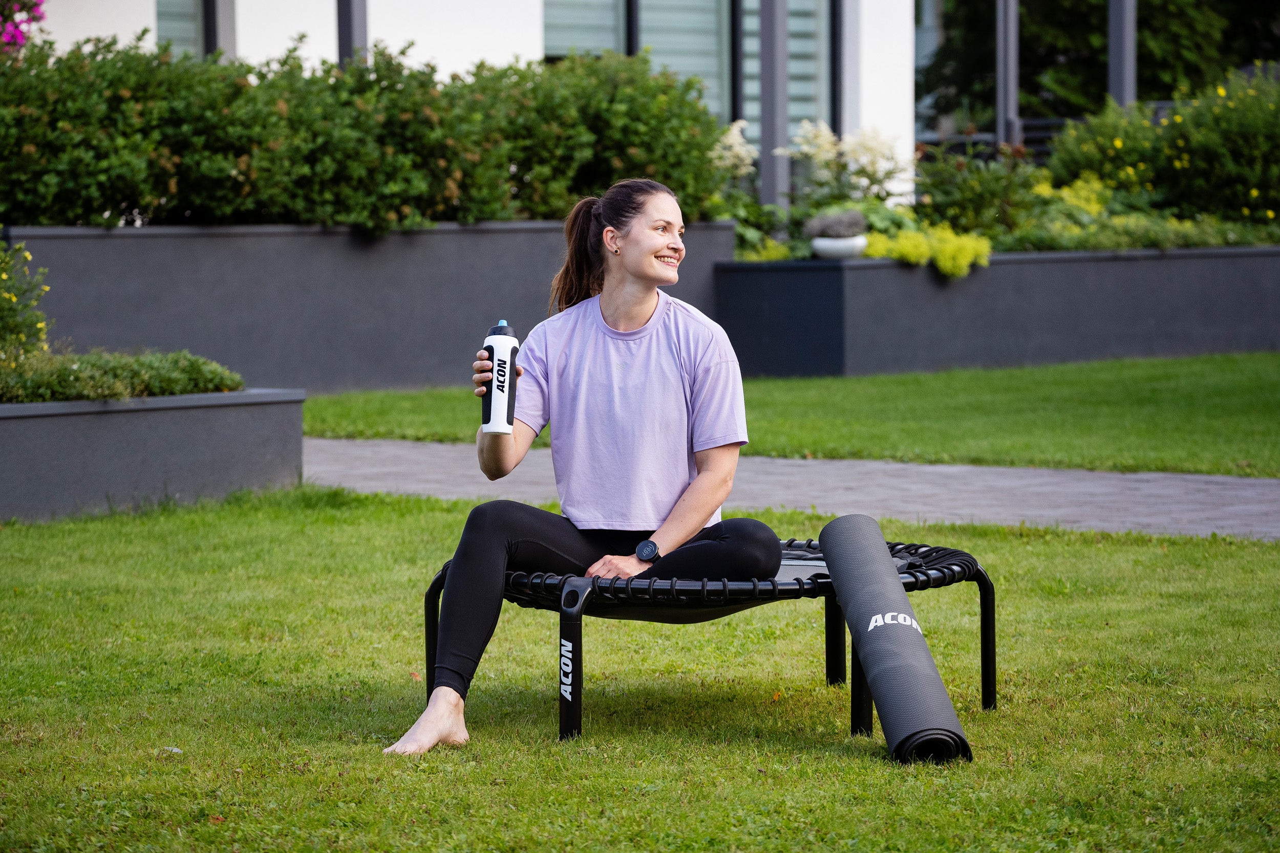 Nainen istuu Aconin juomapullo kädessä fitness-trampoliinilla jumppamaton nojatessa trampoliiniin.