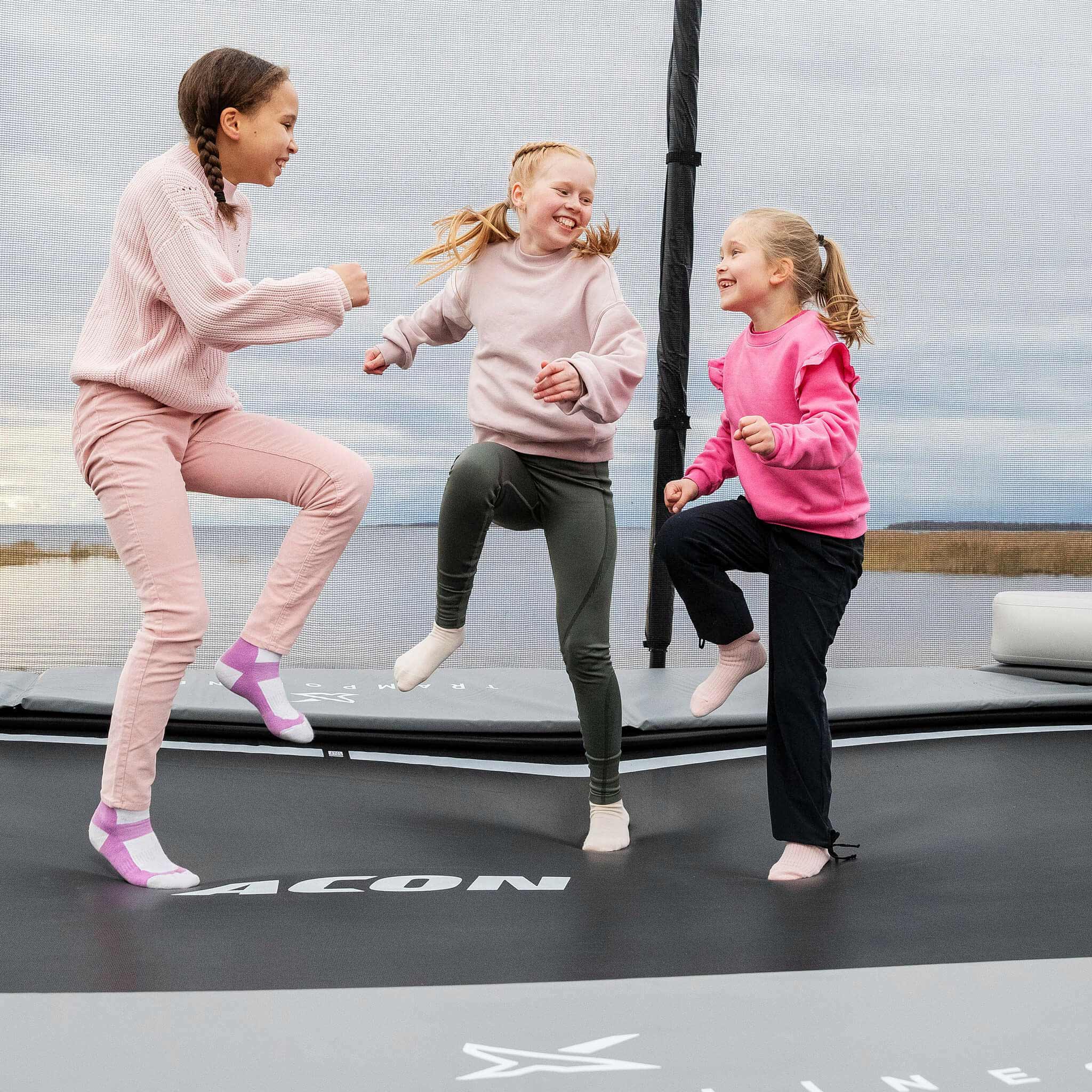 Kolme iloista tyttöä vaaleanpunaisissa paidoissa astelee Acon X -trampoliinilla.