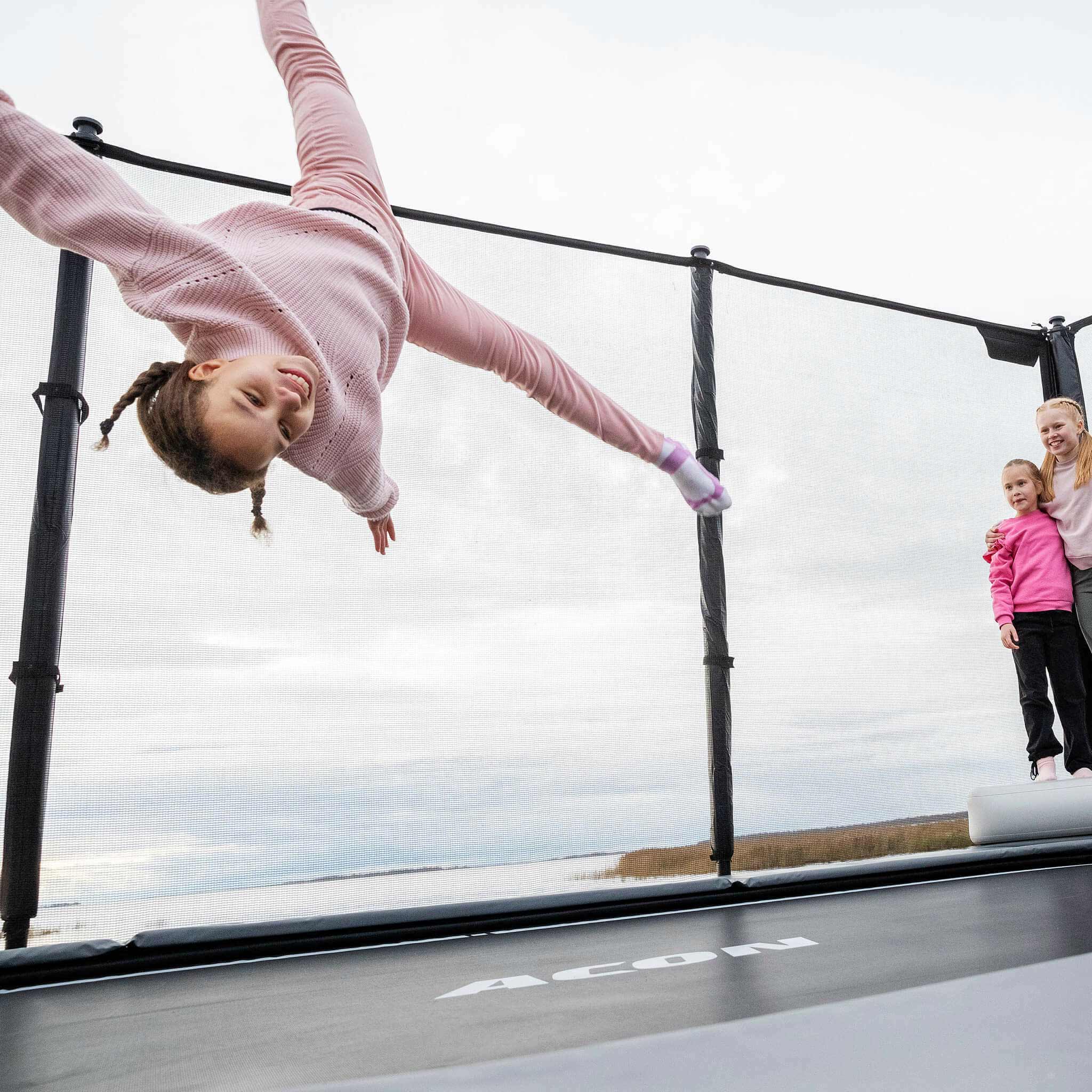 Tyttö hyppää Acon X -trampoliinilla, kaksi tyttöä seisoo ja katsoo.