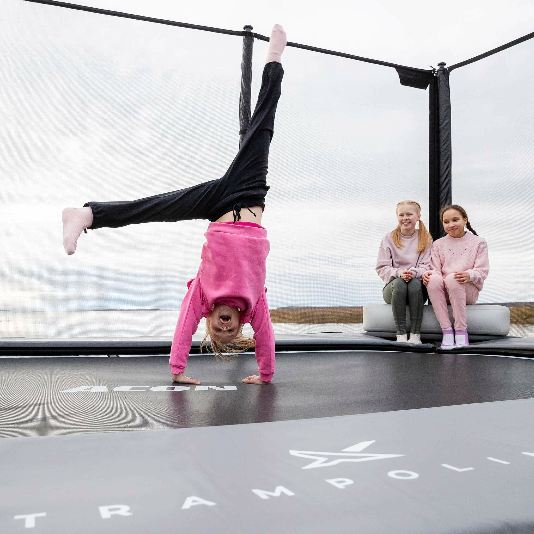 Kaksi tyttöä istuu Acon X Inflatable Triangle Block -puhallettavan kolmioblokin päällä. Tyttö seisoo käsillään trampoliinin päällä.