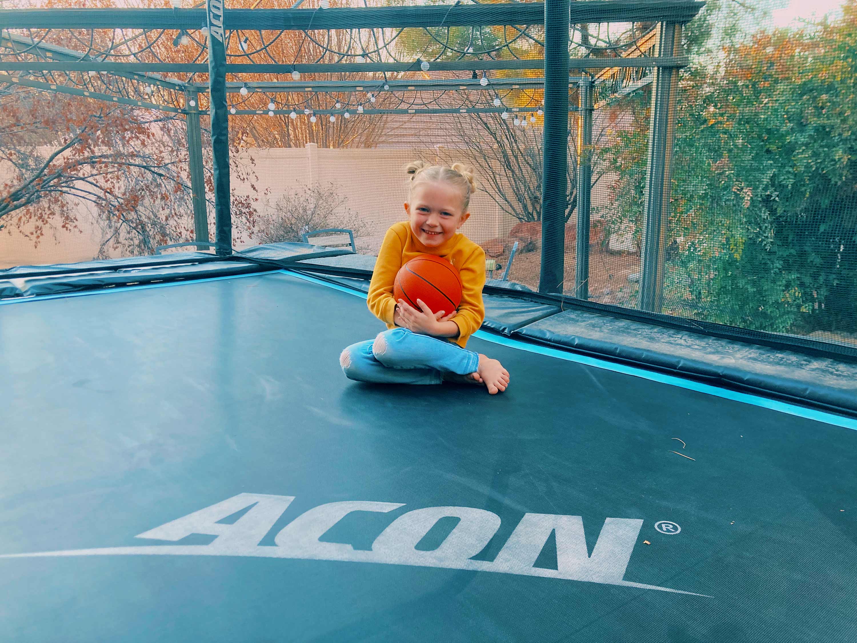 Iloinen tyttö istuu trampoliinilla ja pitelee koripalloa sylissään