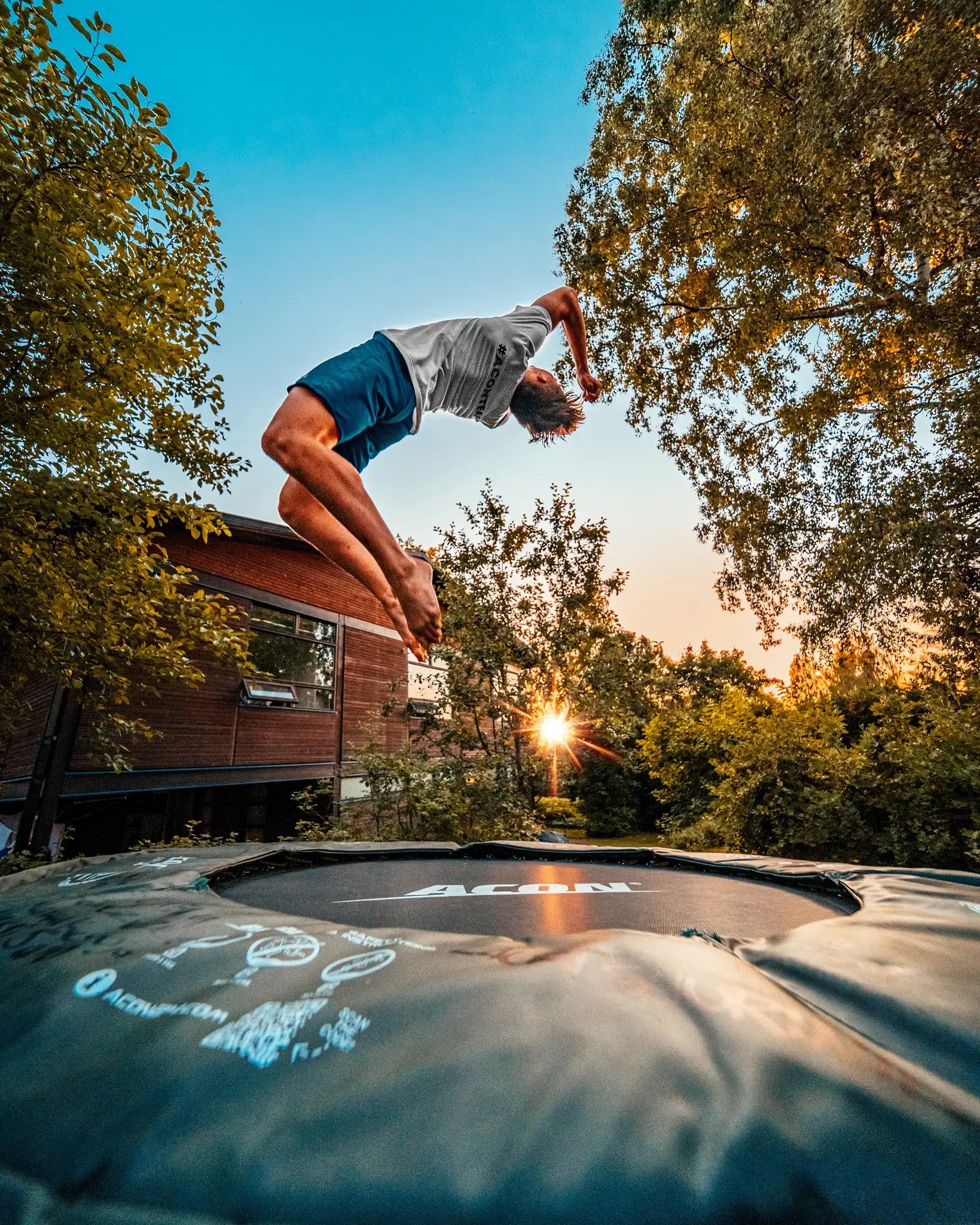 Henkilö tekemässä takaperinvolttia Acon-trampoliinilla pihalla auringonlaskun aikaan.