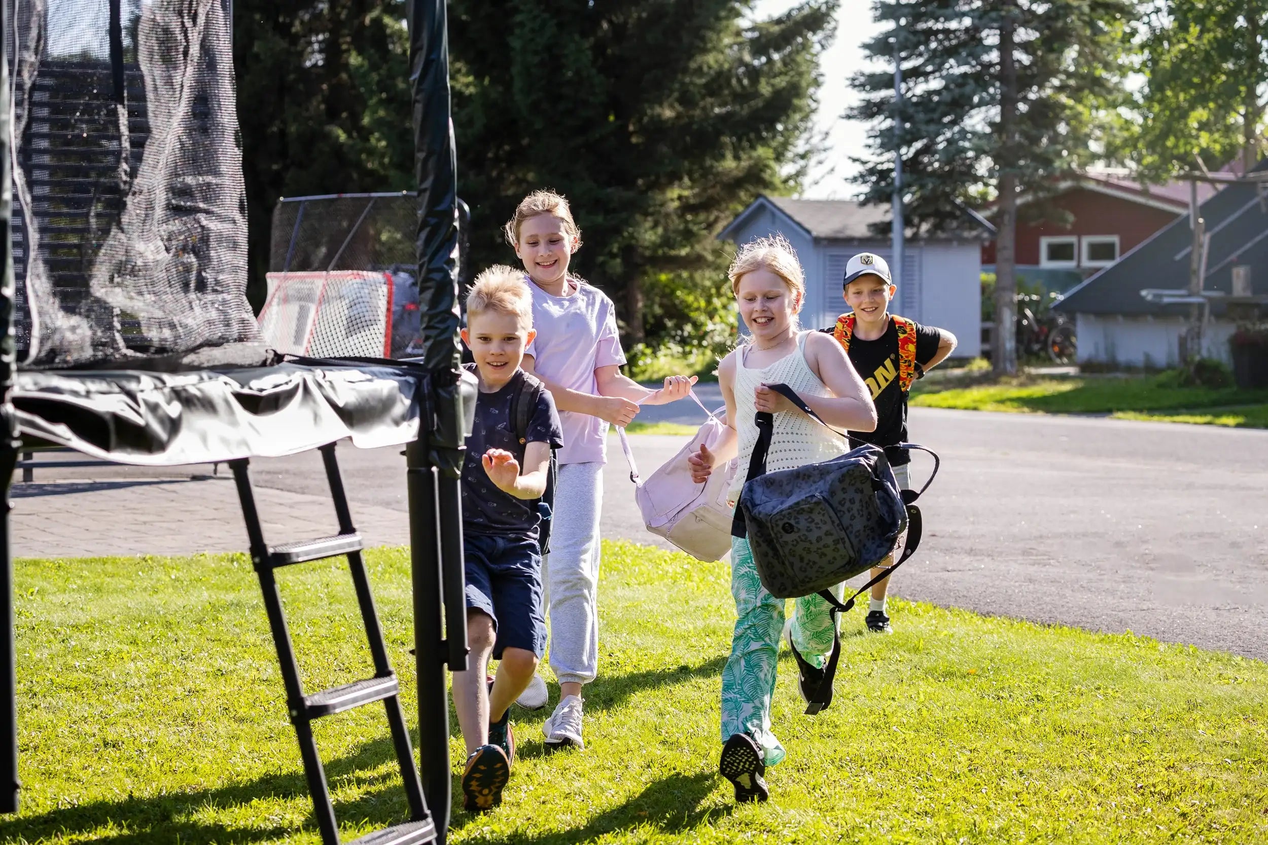 Lapset juoksevat trampoliinille koulun jälkeen. Heillä on mukana reput ja he ovat iloisia.