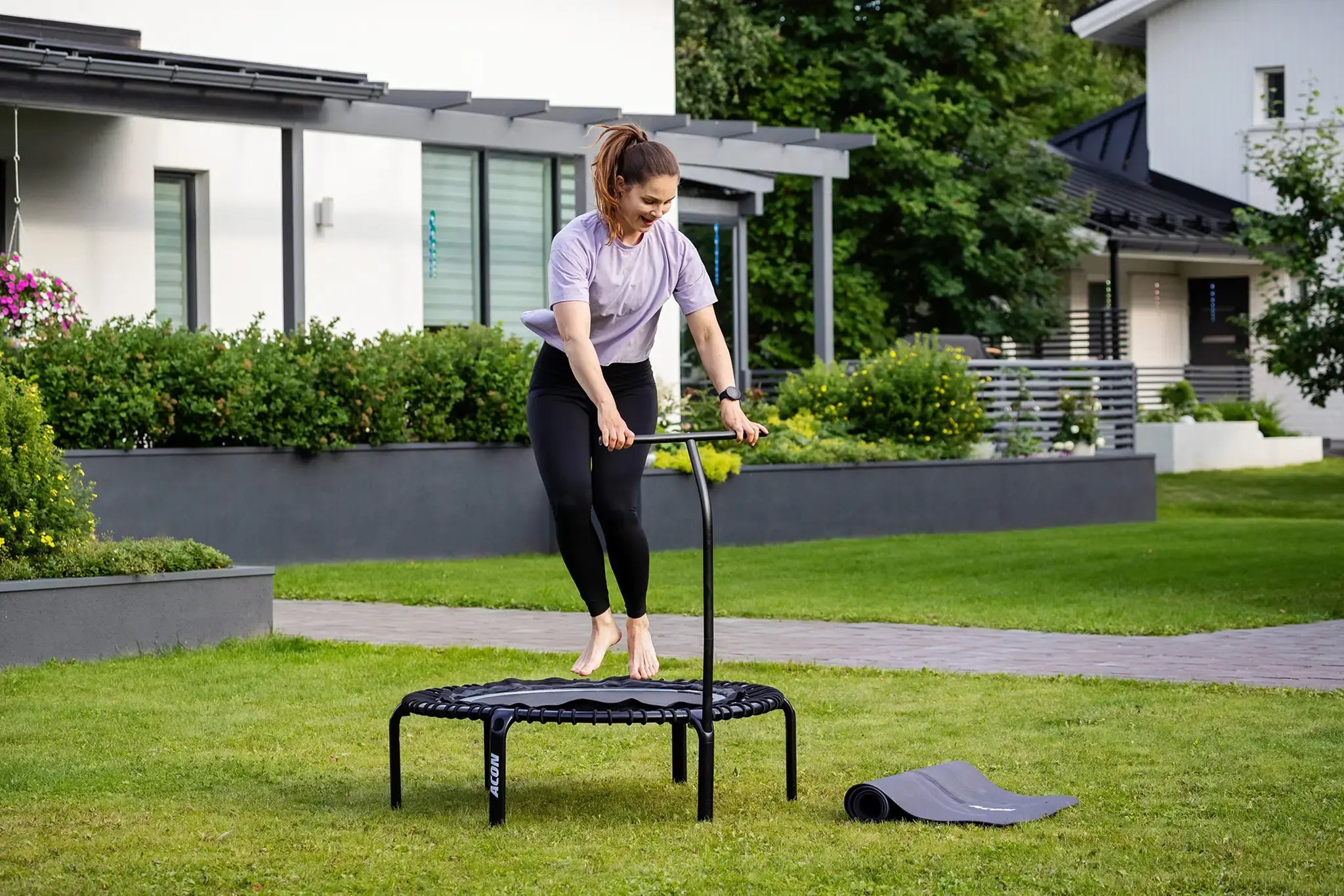 Nainen hyppii aconin fitness-trampoliinilla ulkona nurmikolla, taustalla moderni omakotitalo. 