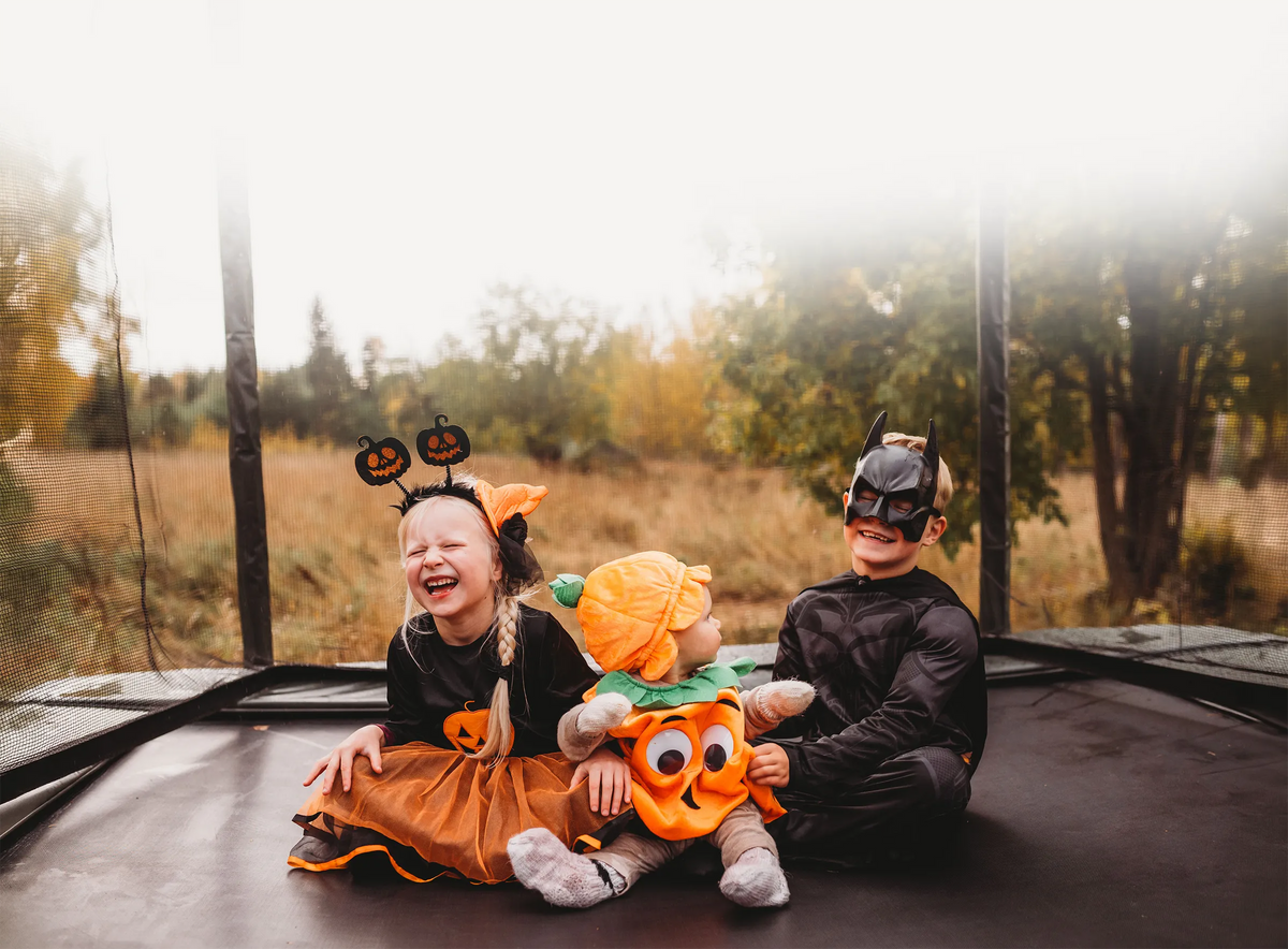 Kolme Halloween-asuihin pukeutunutta lasta istuu yhdessä ACON 16HD -trampoliinilla.