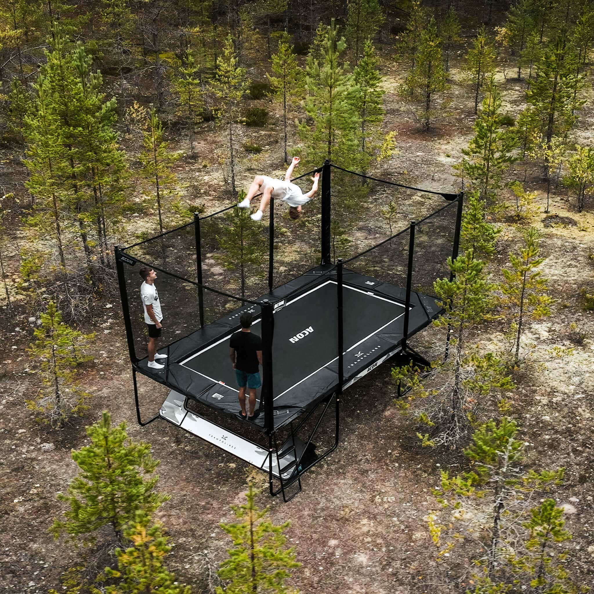 Kolme trampoliinitrikkailijaa Acon X -trampoliinilla pohjoismaisessa mäntymetsässä.