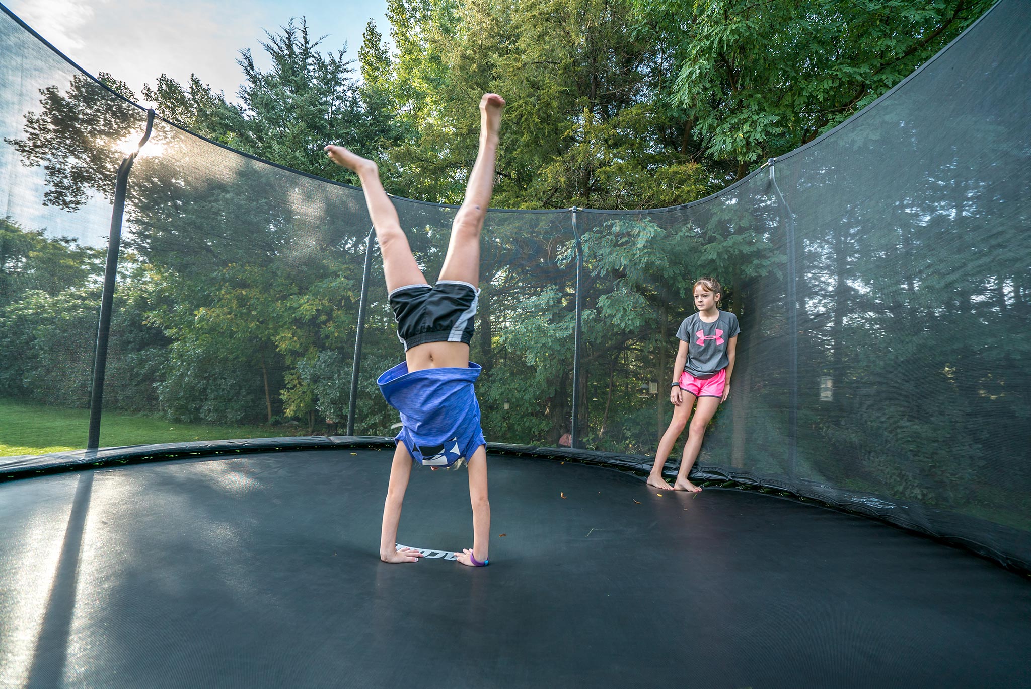 Tyttö seisoo käsillään trampoliinilla, toinen katselee ja nojaa turvaverkkoon
