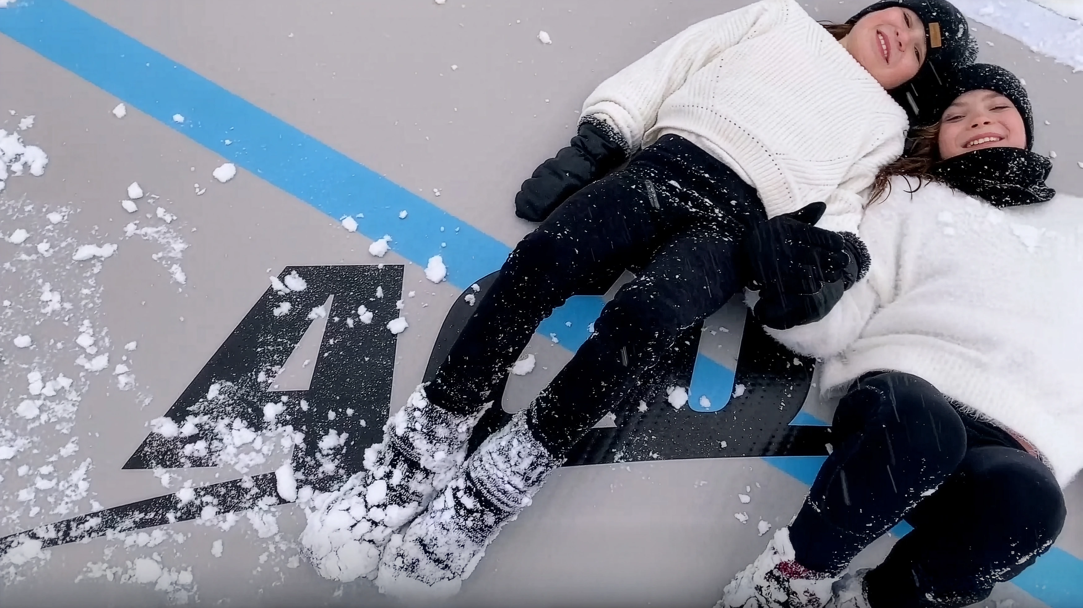 Kaksi hymyilevää sisarusta makaa vierekkäin Acon airtrackin päällä, jonka pinnalla on hieman lunta.