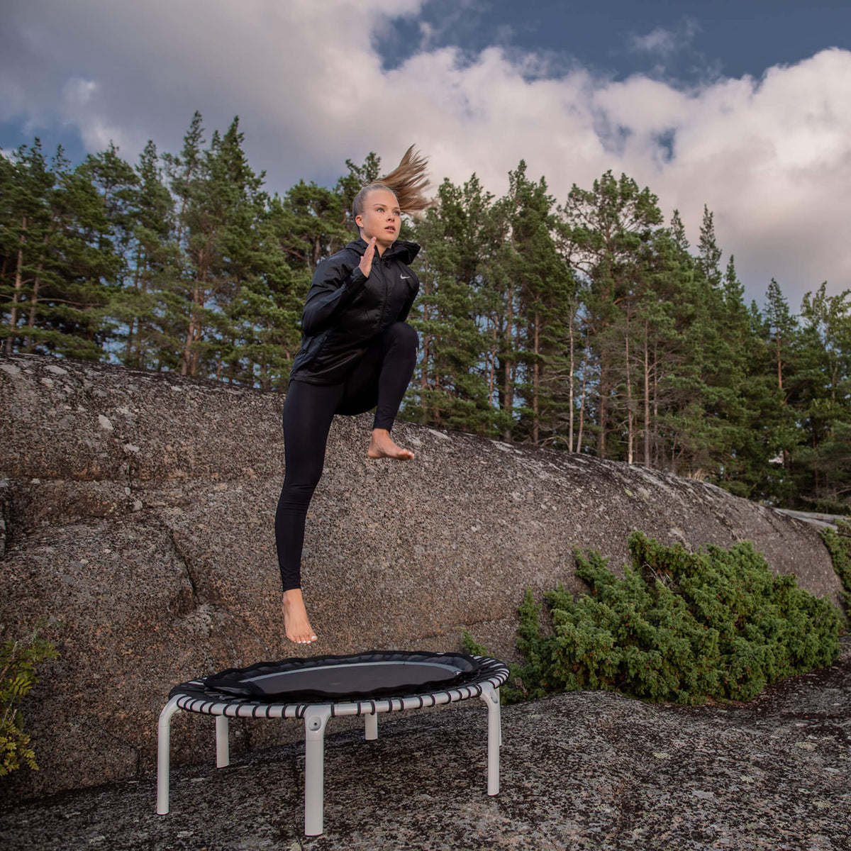 Nainen hyppii valkoisella Acon FIT-trampoliinilla pohjoismaisessa metsämaisemassa.
