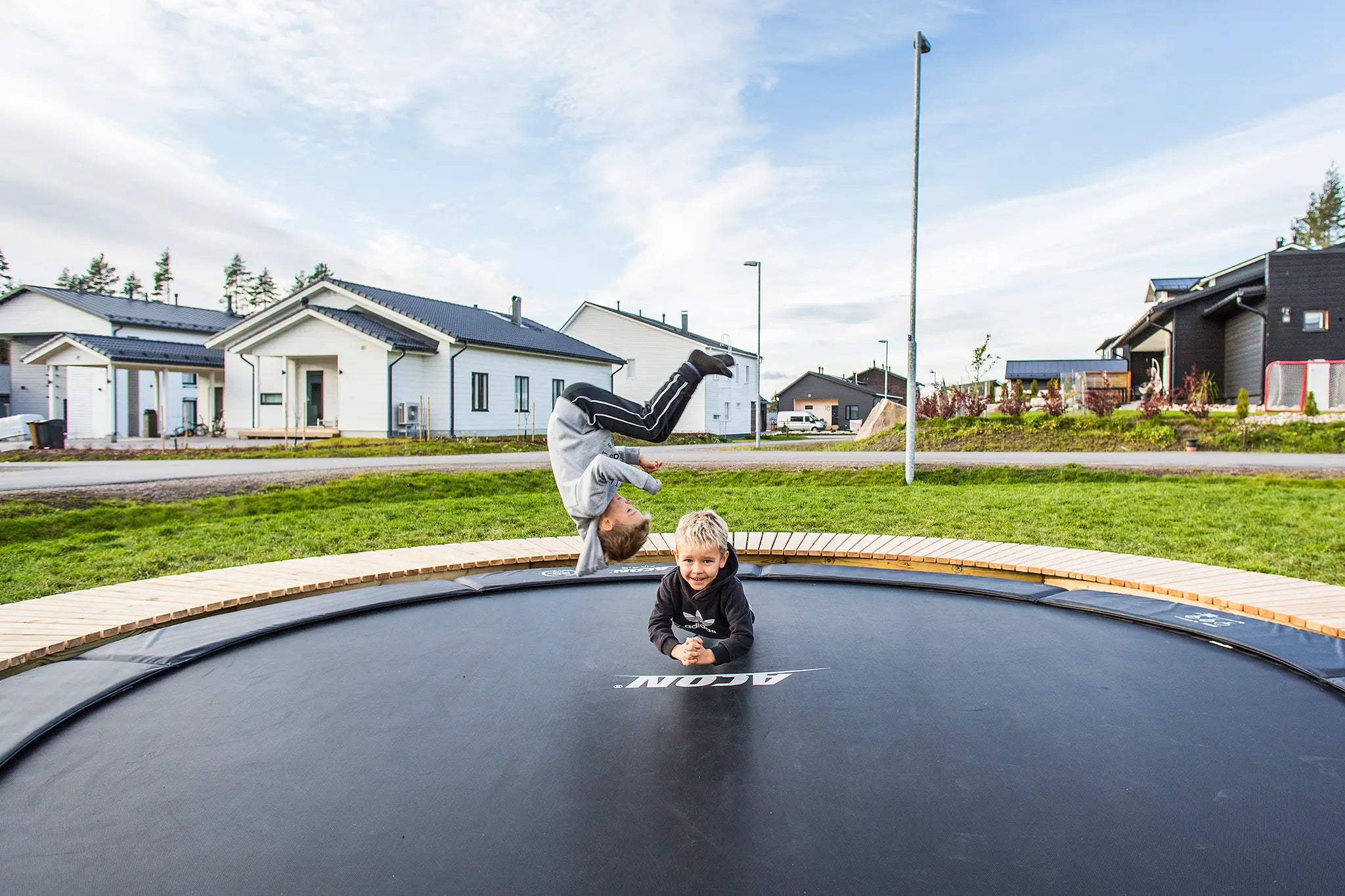 Kaksi pientä poikaa Aconin maahan upotetulla trampoliinilla, toinen makaa, kun toinen tekee tempun. 