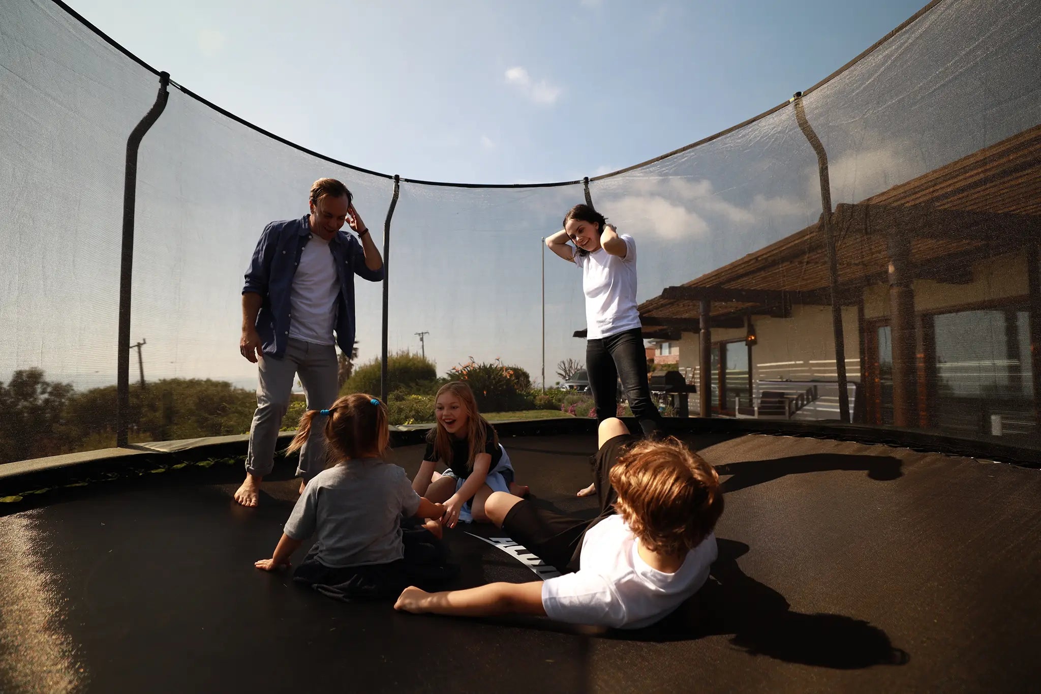 Kaksi aikuista ja kolme lasta Aconin pyöreällä trampoliinilla, jossa on turvaverkko. 