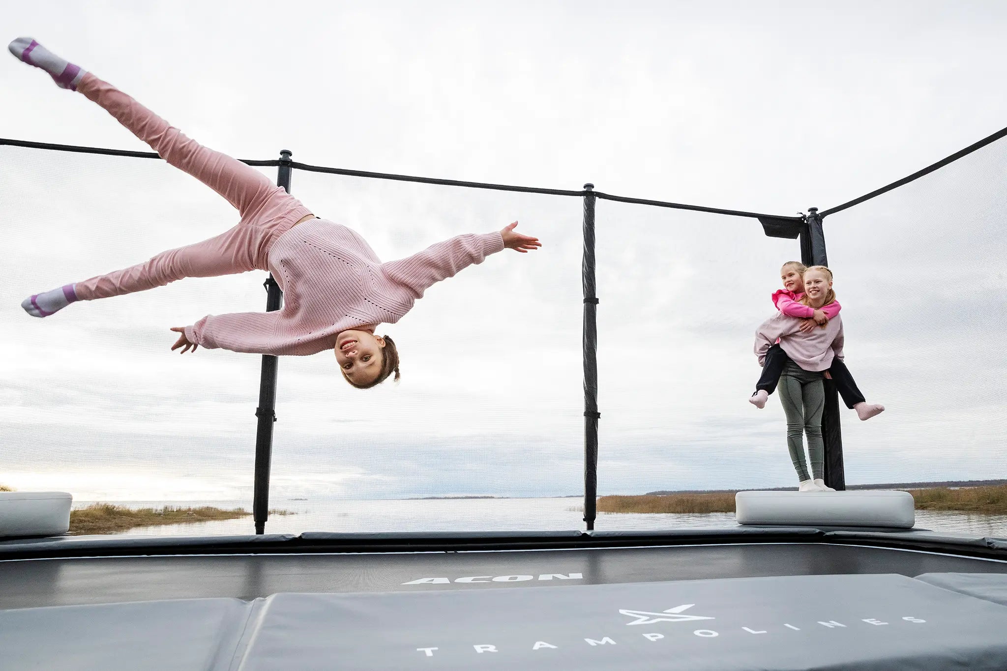 Kolme tyttöä Acon X trampoliinilla, yksi tekee tempun ilmassa, kun kaksi muuta katsovat sivusta. 