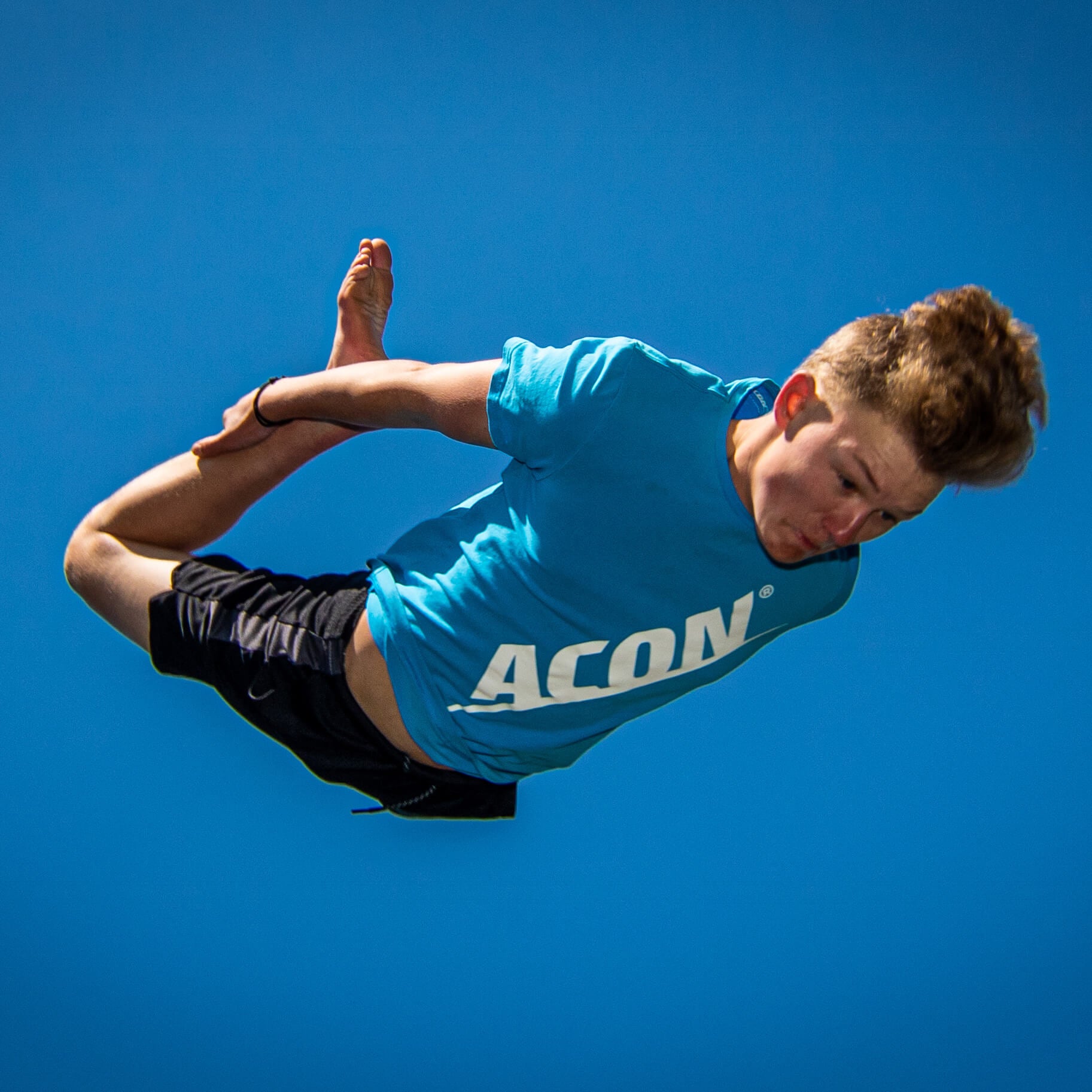 Poika sinisessä ACON t-paidassa hyppää trampoliinilla.
