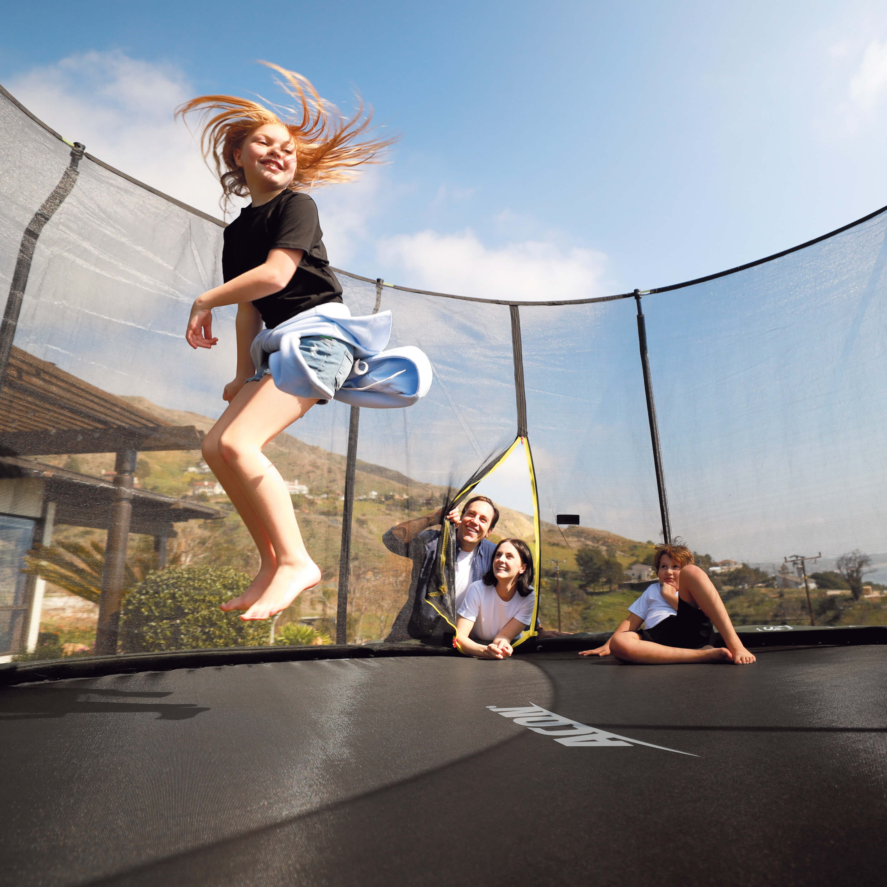 Tyttö hyppää Acon-trampoliinilla, perhe katselee.