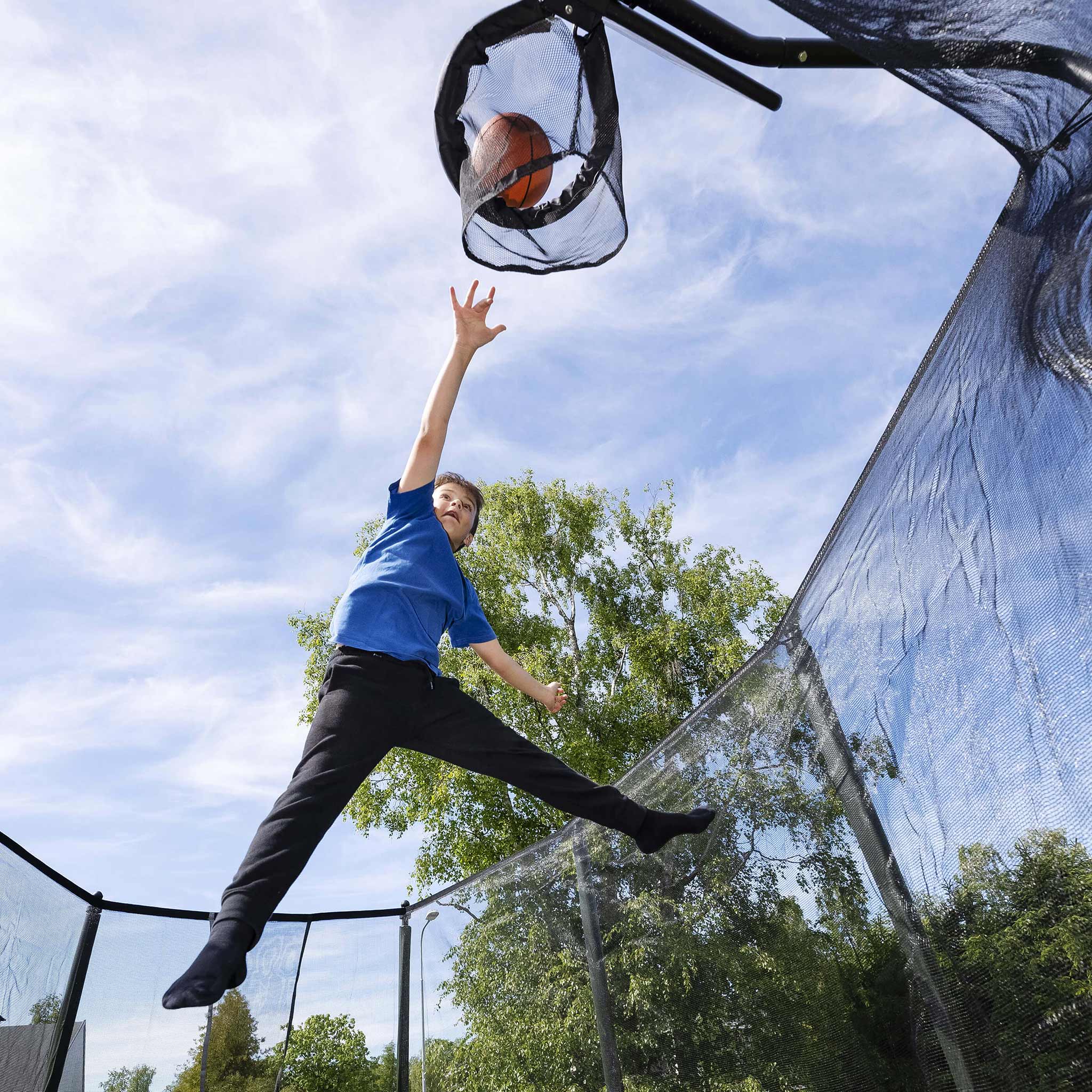 Poika heittää koripallon trampoliinin koriverkkoon.